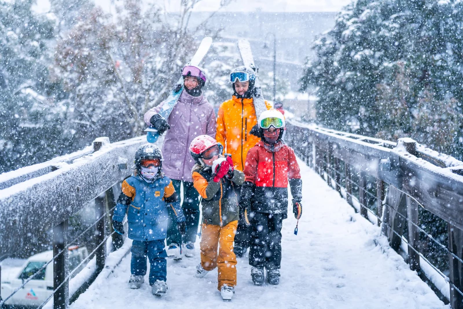 A family carrying skis walks across a village footbridge in falling snow